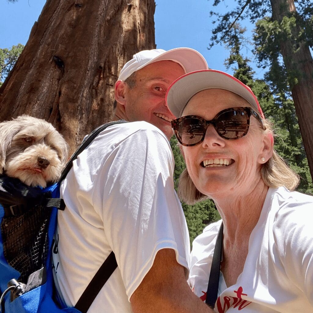 A man and woman with a dog in a backpack with large trees in background