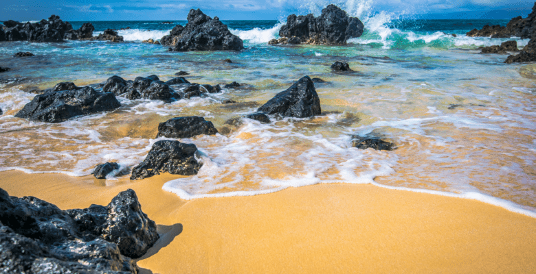 Ocean Waves crashing against black lava rocks with yellow sand in Maui, Hawaii