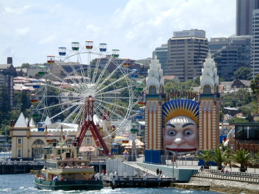 small carnival area with ferris wheel along a river with a boat passing by