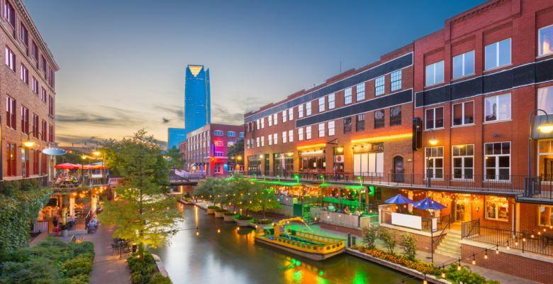 A city view of a skyscraper and river walk with restaurants along the side of it in Oklahoma City.
