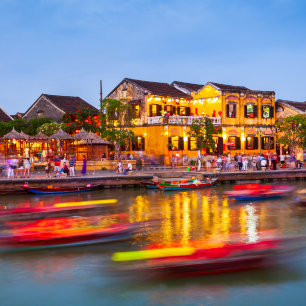 Boats passing through a canal in the city of Hoi An at dusk with restaurants, stores, and people walking next to it.