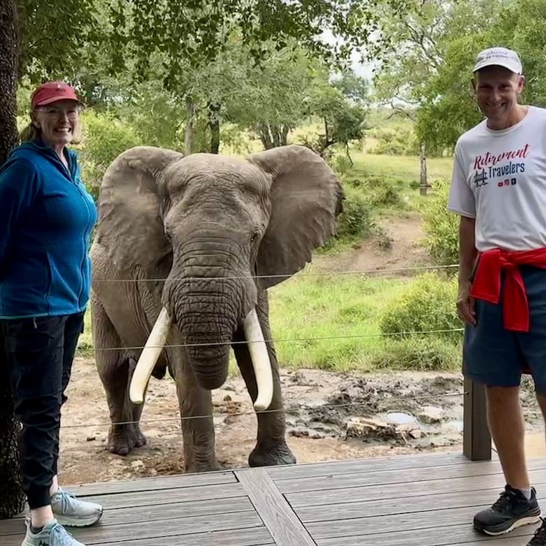 John & Bev standing on the deck of the lodge with an elephant in the background.